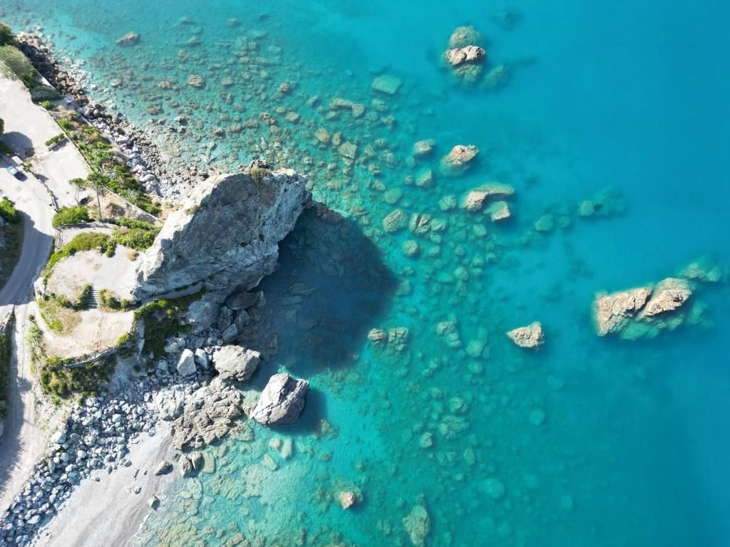 An aerial view of a coastal area with clear turquoise waters, rocky outcrops, and a sandy beach.