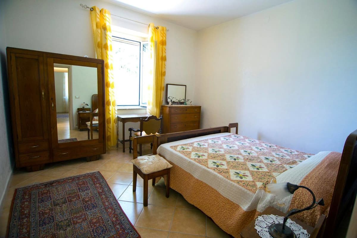 Een slaapkamer met een bed, een bureau, een stoel, een kledingkast en een raam met gordijnen.