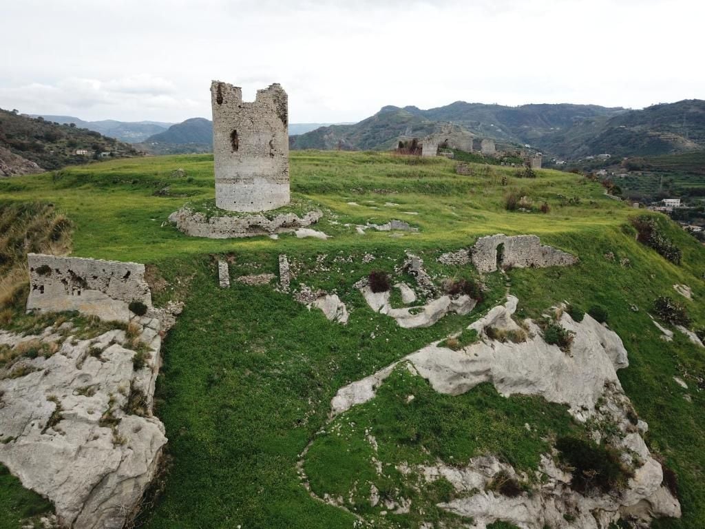 Eine historische Burgruine mit einem zentralen Turm, umgeben von grüner Landschaft und Hügeln im Hintergrund.