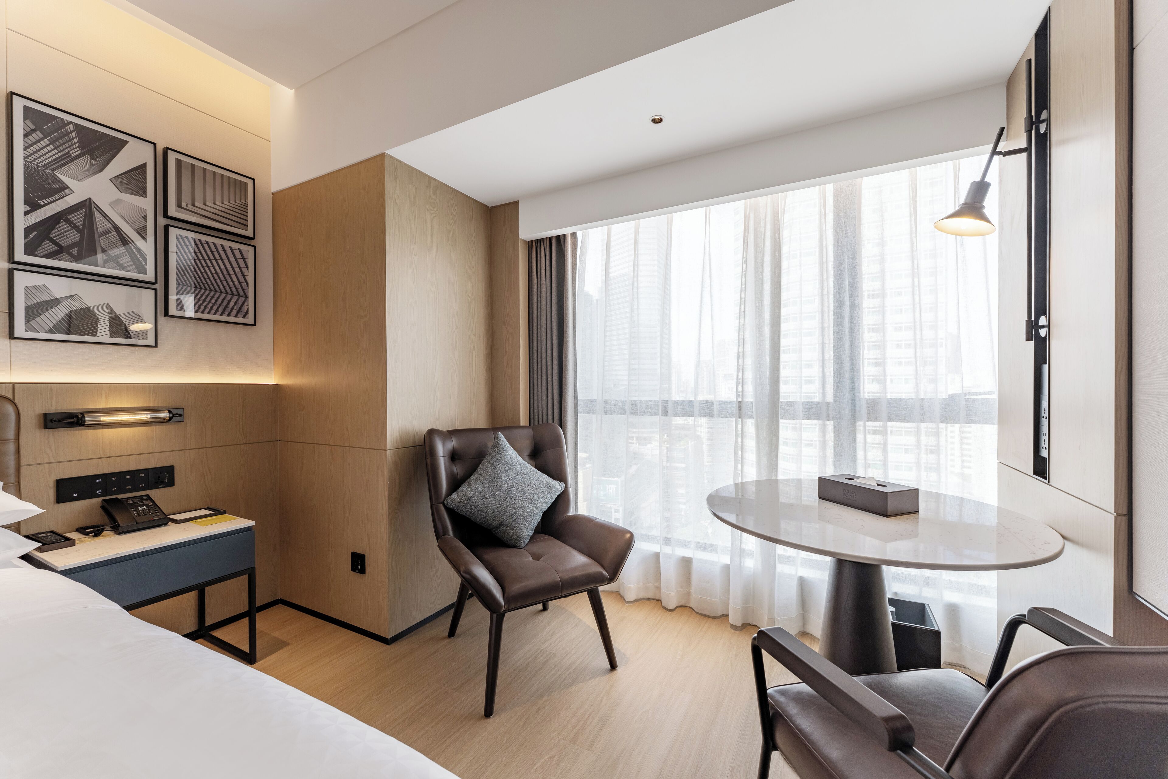 Een moderne hotelkamer met een groot raam, een ronde tafel, een comfortabele stoel, een bureau met telefoon en kunst aan de muur.