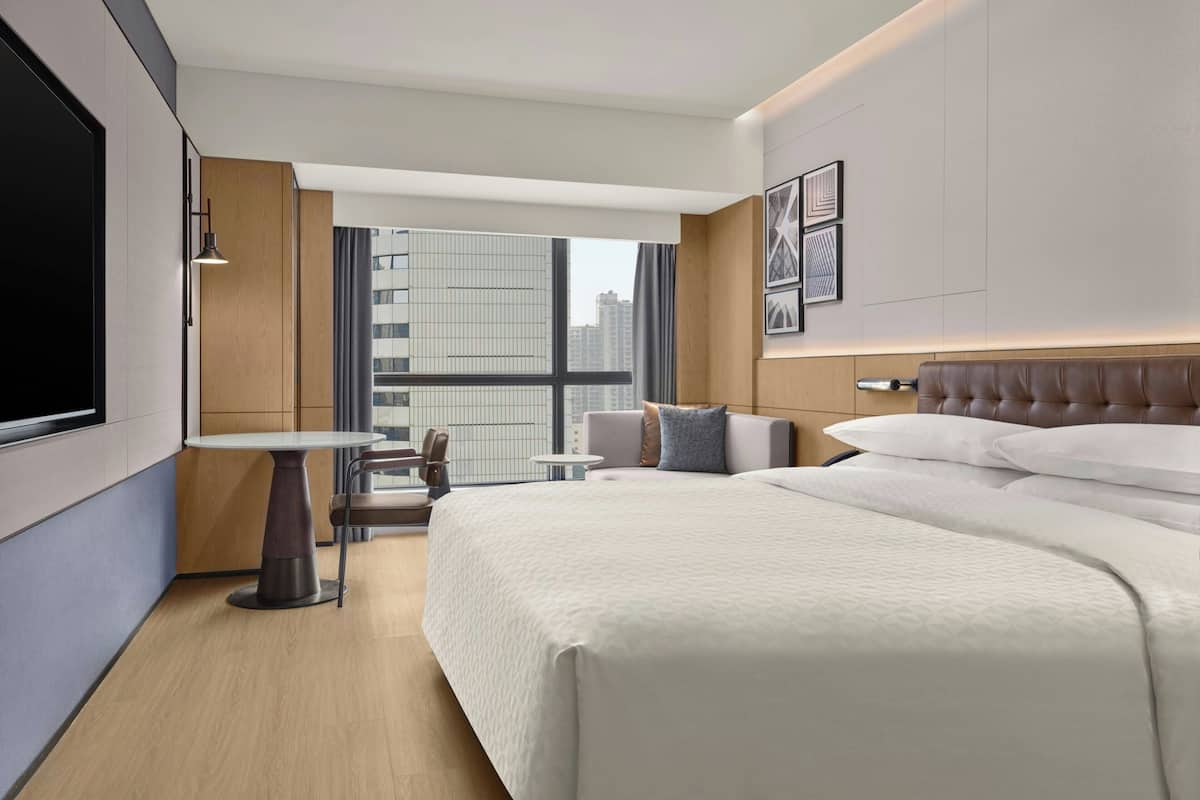 Ein Hotelzimmer mit einem großen Bett, einem kleinen Tisch, einem Sessel, einem Fernseher und einem Fenster mit Blick auf die Stadt.