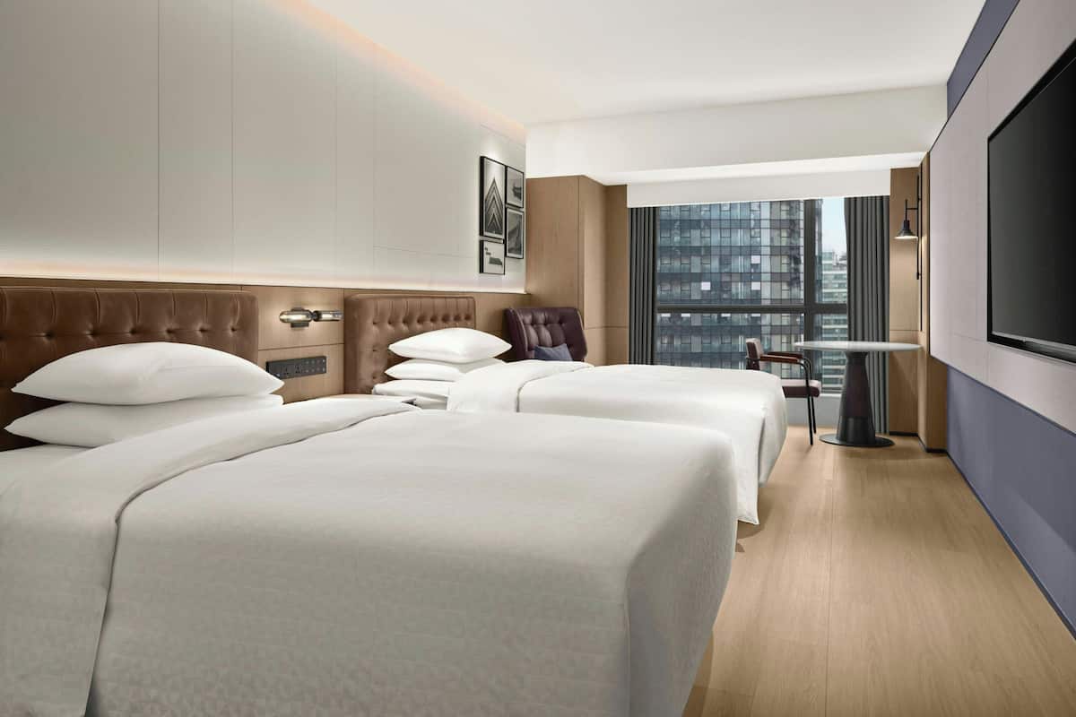 Ein Hotelzimmer mit zwei Betten, einem Fernseher, einem kleinen Tisch und Blick auf die Stadt.