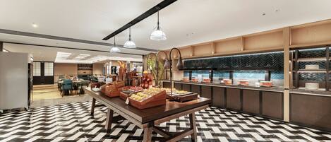A modern hotel lobby with a checkered floor, a buffet table with various food items, and a seating area in the background.