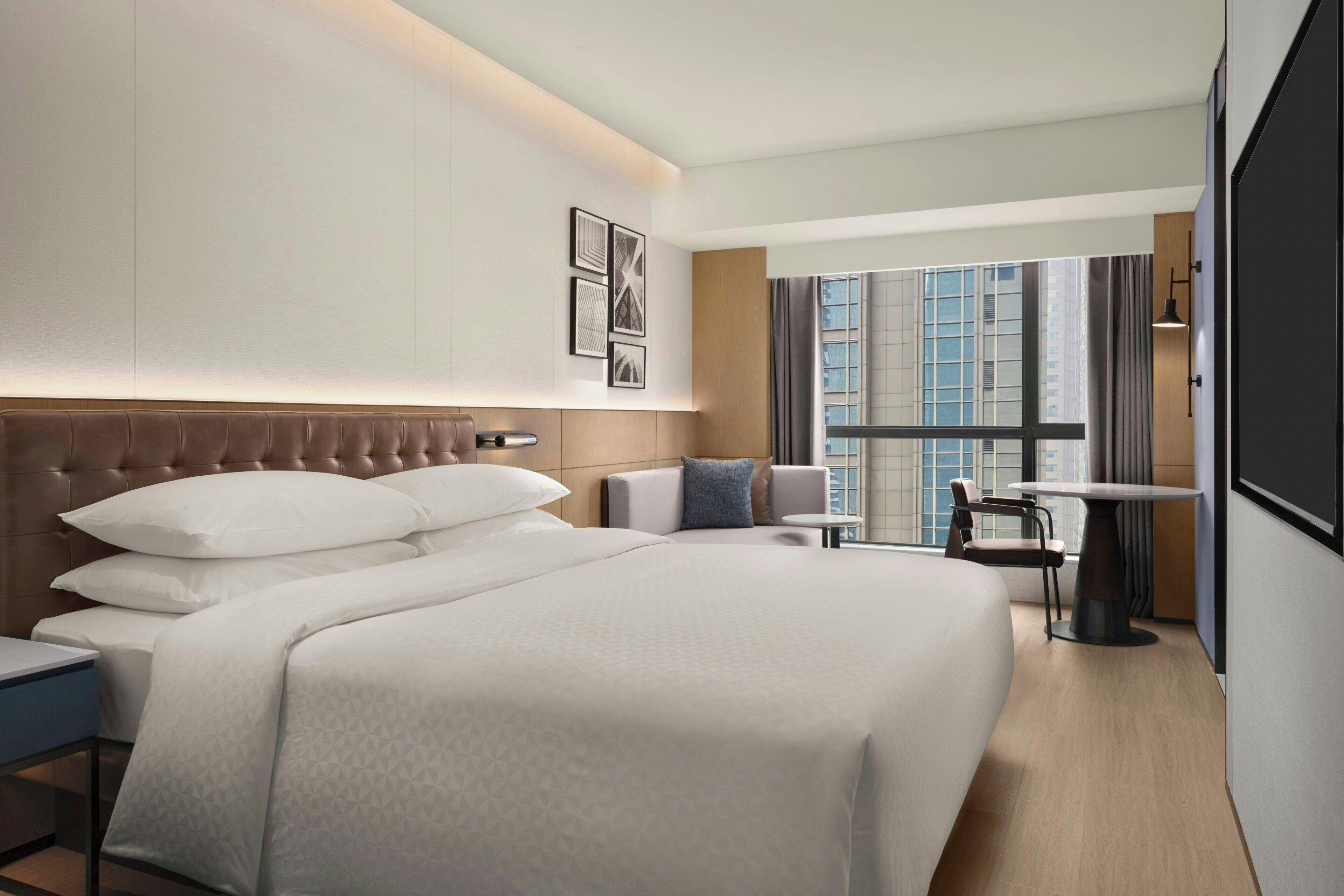 A hotel room with a large bed, a small table, a chair, and a window with a city view.