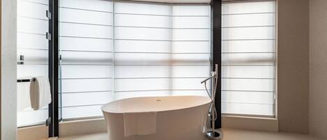 A corner bathroom with a freestanding bathtub, large windows with blinds, and a towel rack.