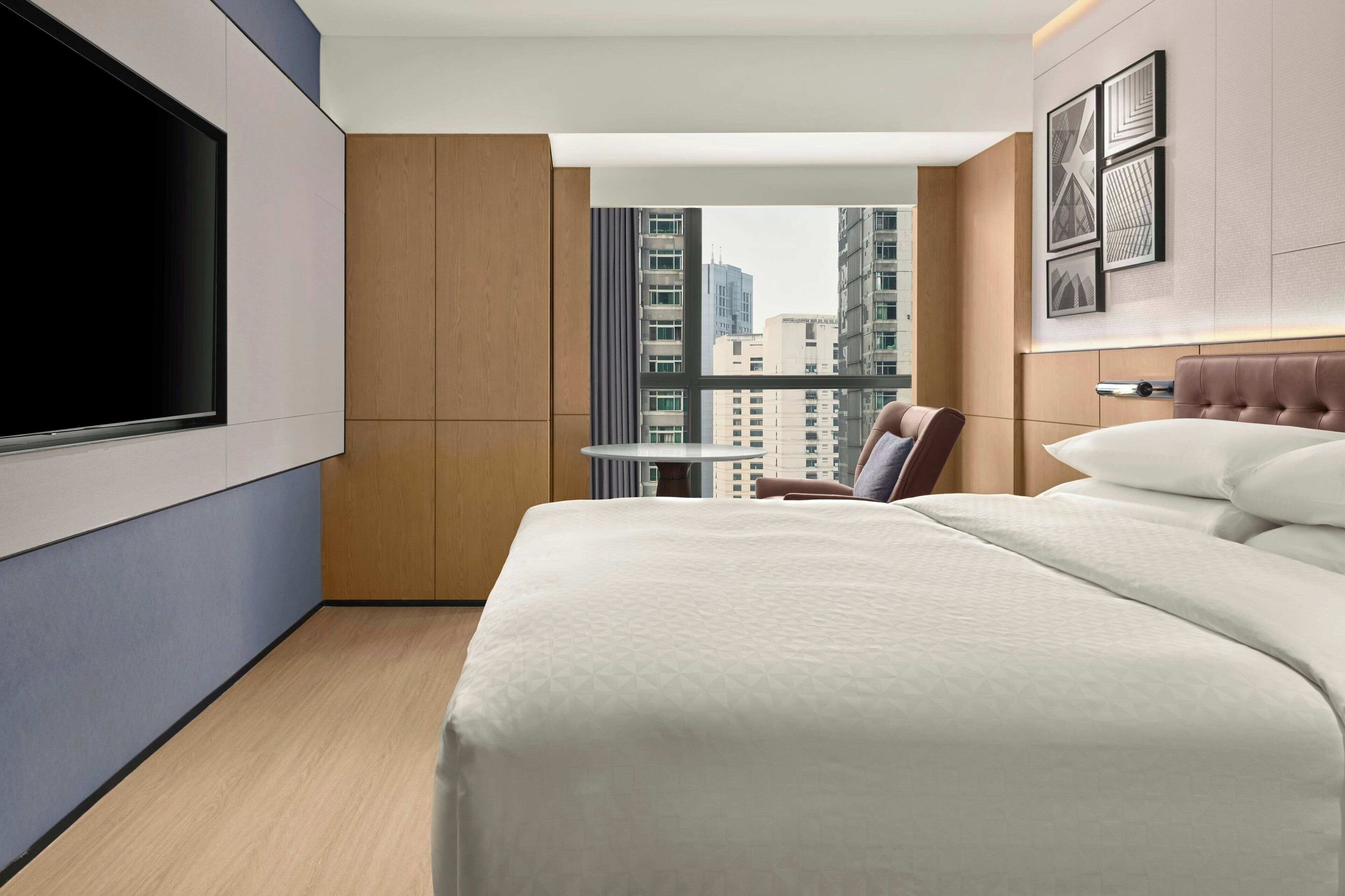 Quarto de hotel moderno com uma cama grande, televisão, mesa de jantar e vista para a cidade.