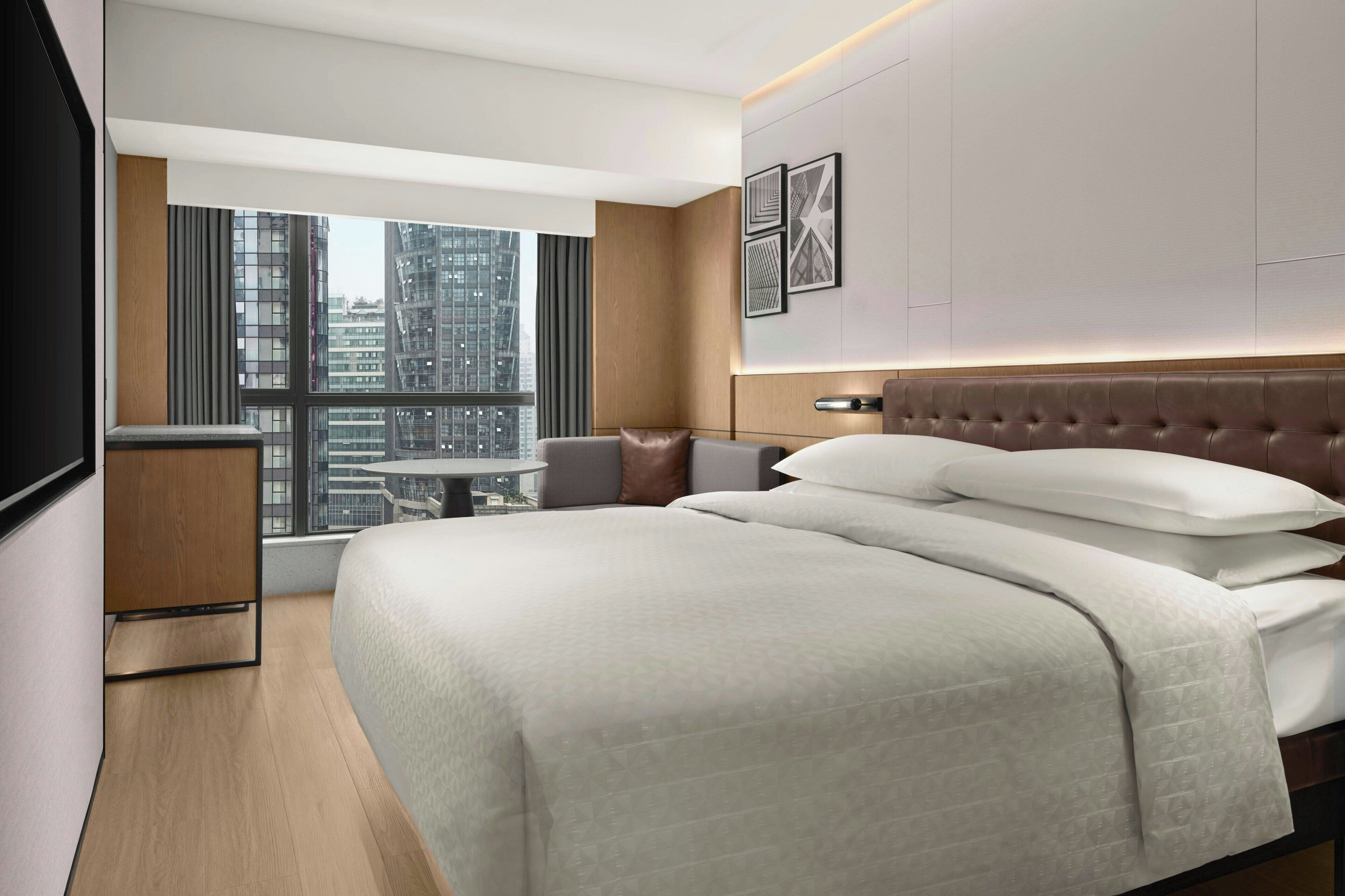A modern hotel room with a large bed, a small seating area, a city view, and a flat-screen TV.