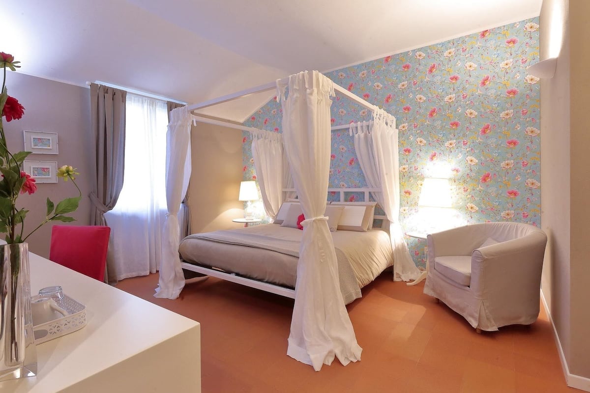 Een slaapkamer met een hemelbed, nachtkastje, stoel en bloemenbehang.