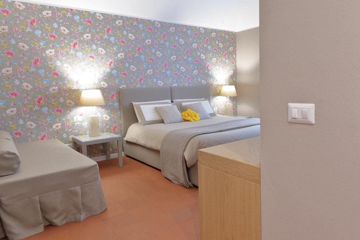 Een slaapkamer met bloemenbehang, een bed met kussens, een nachtkastje met een lamp en een nachttafel.