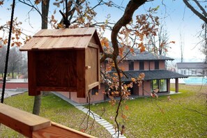 A wooden birdhouse attached to a tree, a wooden bench, and a building with a tiled roof in the background. - Agriturismo Relais Tenuta il Cigno (Villanterio)