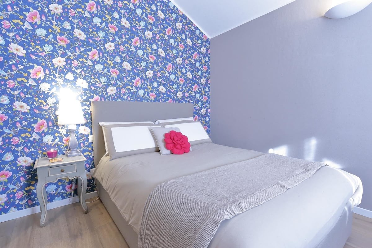 Een slaapkamer met bloemenbehang, een bed met witte beddengoed en een rood kussen, een nachtkastje met een lamp, en een kleine tafel met een kaars.