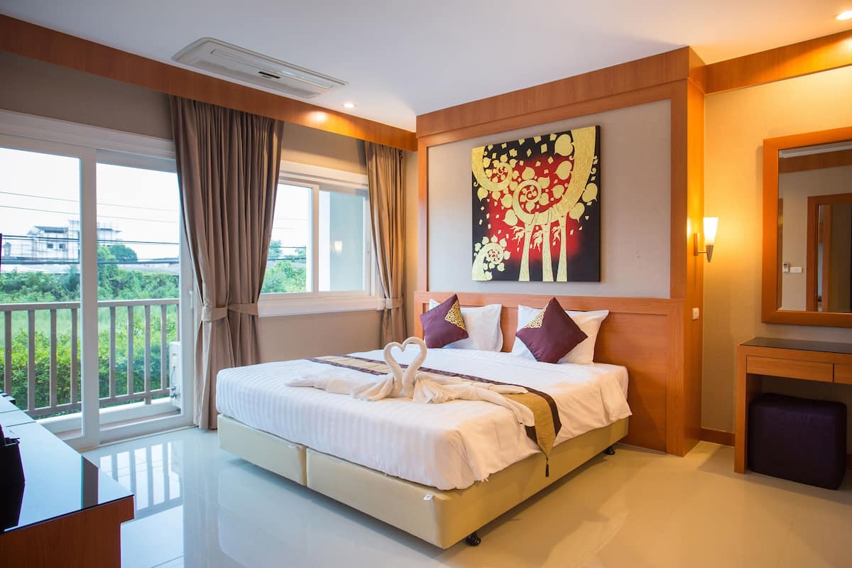 Quarto de hotel com uma cama grande, uma escrivaninha, uma cadeira, janela com cortinas, um quadro na parede e um banquinho pequeno.