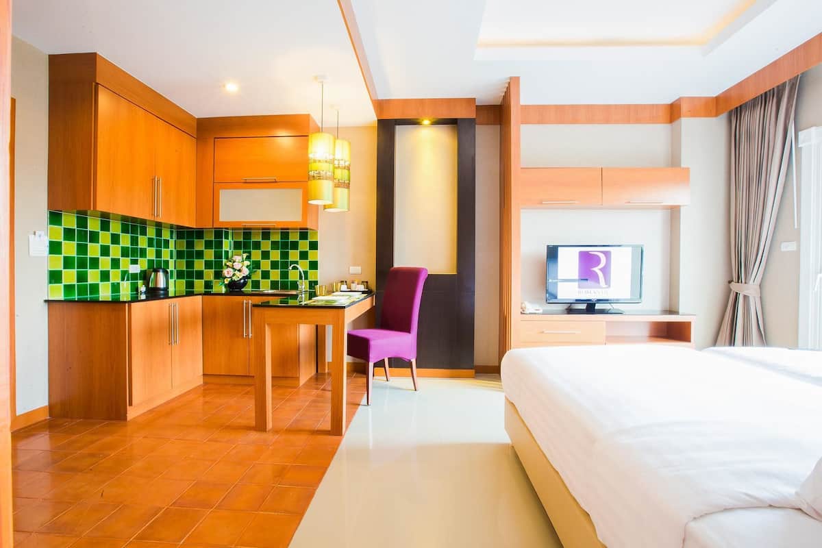 Quarto de hotel com cama, televisão, secretária com cadeira e cozinha com armários de madeira e azulejos verdes.