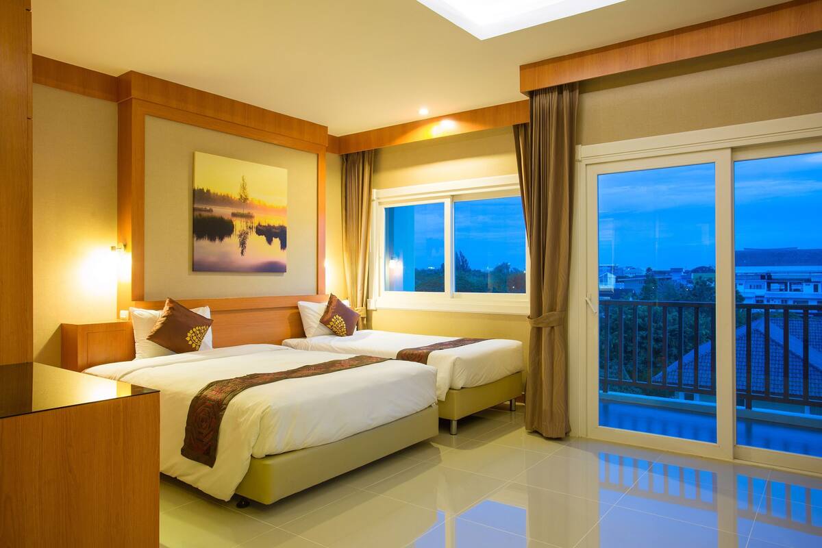 Quarto de hotel com duas camas, um quadro, uma janela grande com cortinas e vista para a cidade.