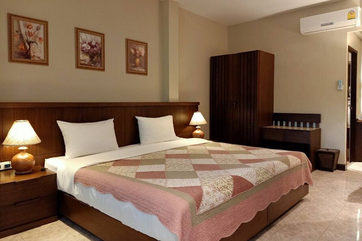 A hotel room with a large bed, wooden headboard, two bedside tables with lamps, a wardrobe, and a wall-mounted air conditioner.