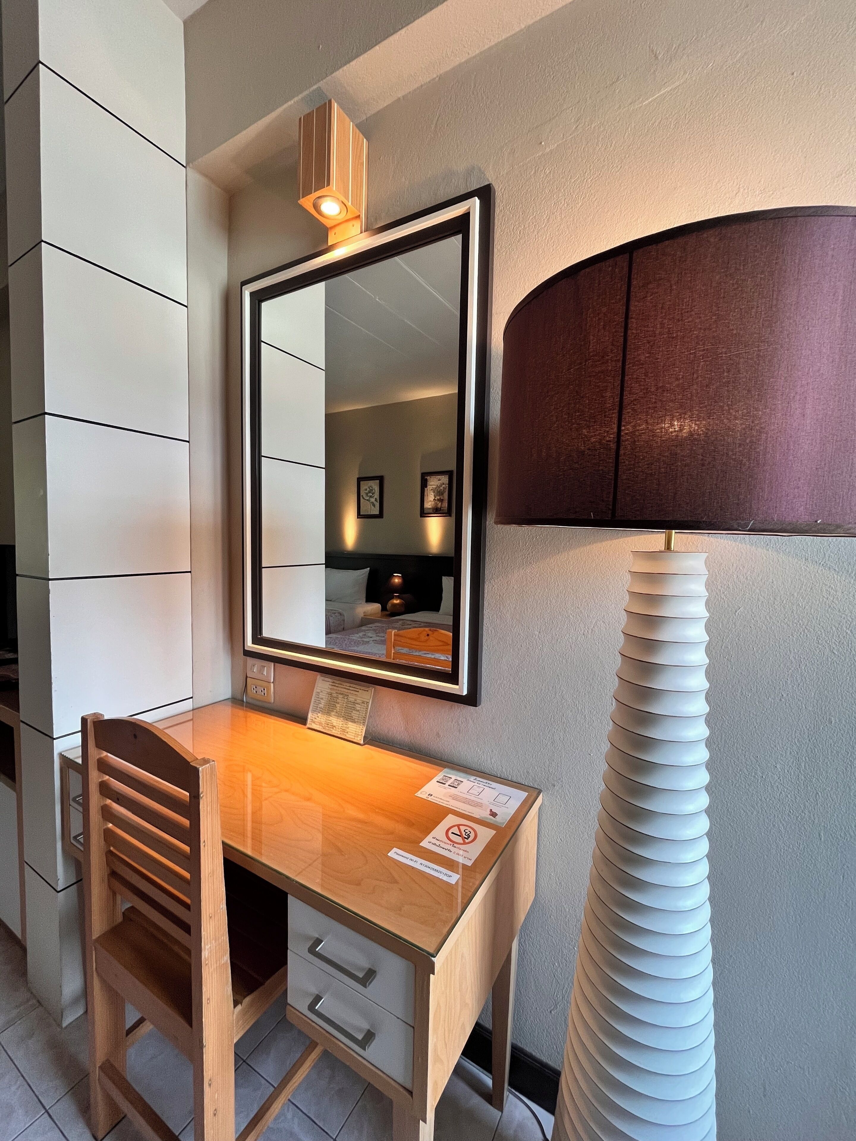A compact hotel room with a wooden desk, a mirror, a lamp, and a wall-mounted shelf.