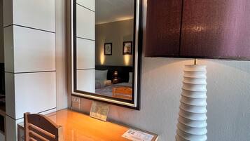 A compact hotel room with a wooden desk, a mirror, a lamp, and a wall-mounted shelf.