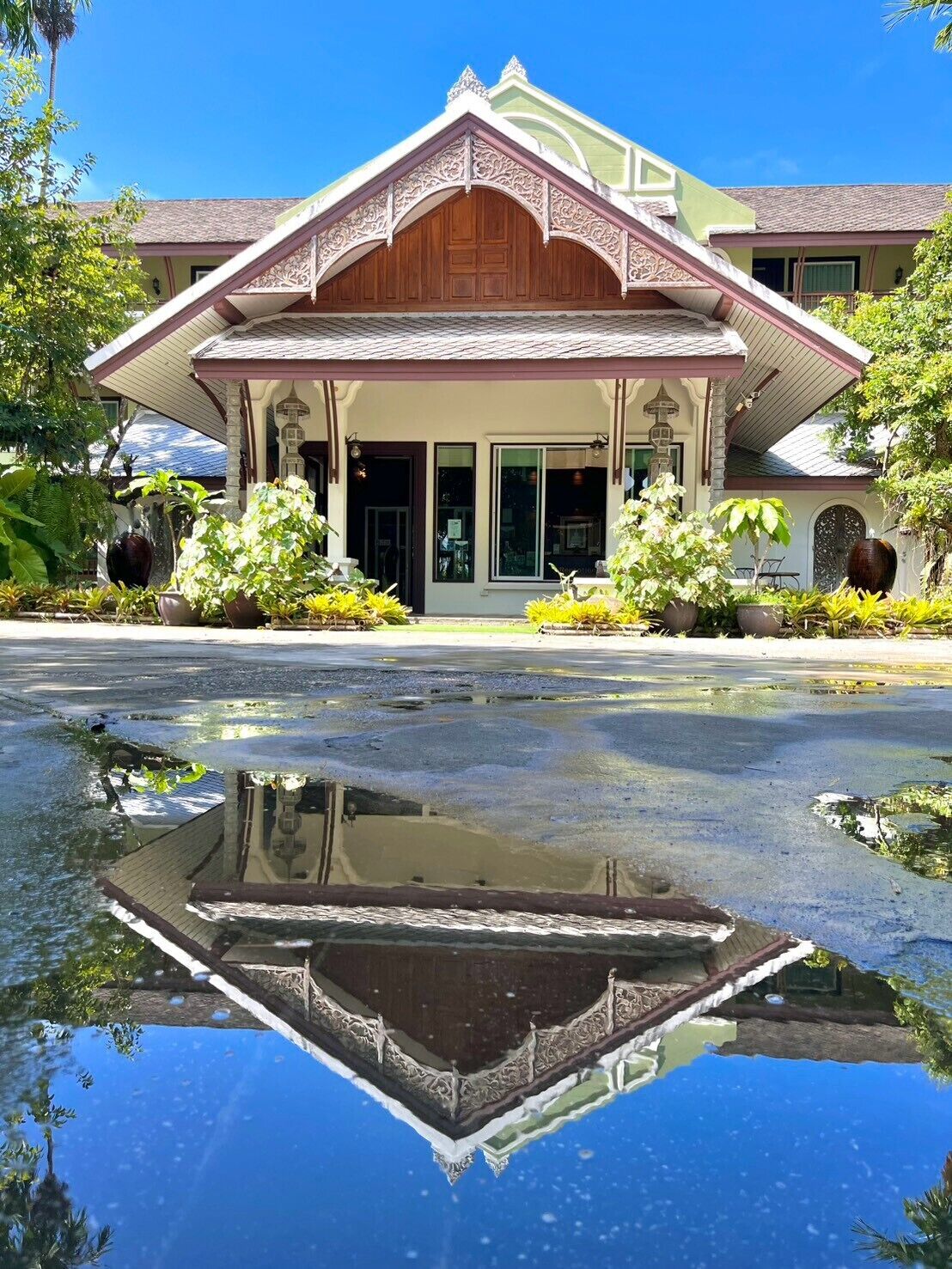 A building with a traditional architectural style, featuring a central wooden structure with ornate detailing, surrounded by lush greenery and a reflective water feature in front.
