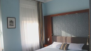 A neatly made bed with white linens and a pillow with stripes, a wooden headboard, a bedside table with a lamp, and a framed picture on the wall. - Hotel Am Roonplatz (Bonn)