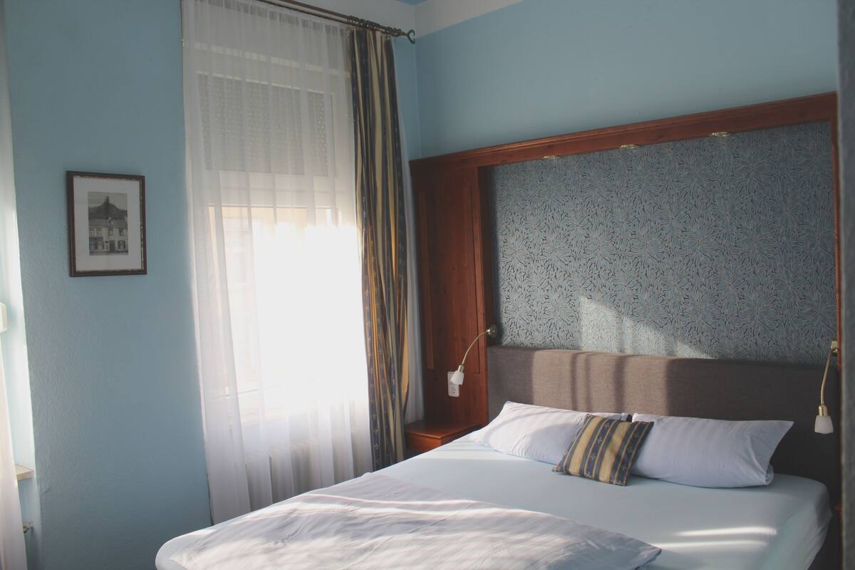 A neatly made bed with white linens and a pillow with stripes, a wooden headboard, a bedside table with a lamp, and a framed picture on the wall.