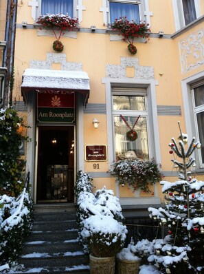 Entrada de un hotel cubierta de nieve, con un cartel que dice "Am Roonplatz" y un árbol de Navidad adornado.