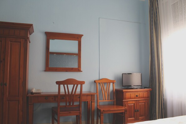Una habitación con un escritorio de madera, dos sillas, un televisor sobre un mueble y un espejo en la pared.