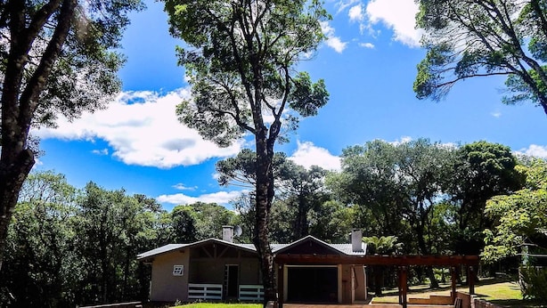 A rustic cabin with a covered porch and a garage, surrounded by lush greenery and a clear blue sky. - Residencial Encantos da Natureza (Canela)