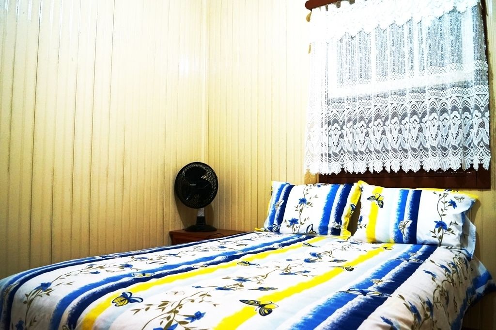 A bedroom with a bed, a bedside table, a fan, and a window with lace curtains.