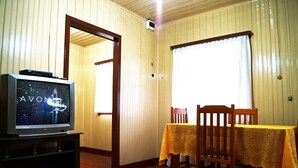 A room with a television displaying a news channel, a dining table with a yellow tablecloth, wooden chairs, and a wooden door leading to another room. - Residencial Recanto do Sossego Gramado (Gramado)