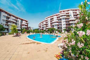 Una piscina envoltada d'edificis d'apartaments amb balcons, cel blau clar i vegetació.