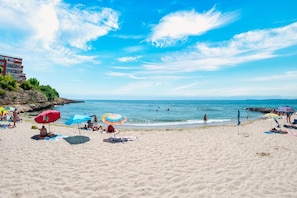 Una platja amb gent prenent el sol i banyant-se, un penya-segat rocos i un cel blau clar.