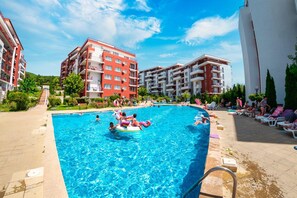 Una piscina rodeada de edificios de varios pisos con balcones, cielo despejado y algunas personas en el agua.