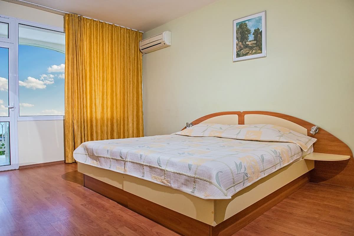 A bedroom with a bed, a nightstand, a window with curtains, and an air conditioning unit.