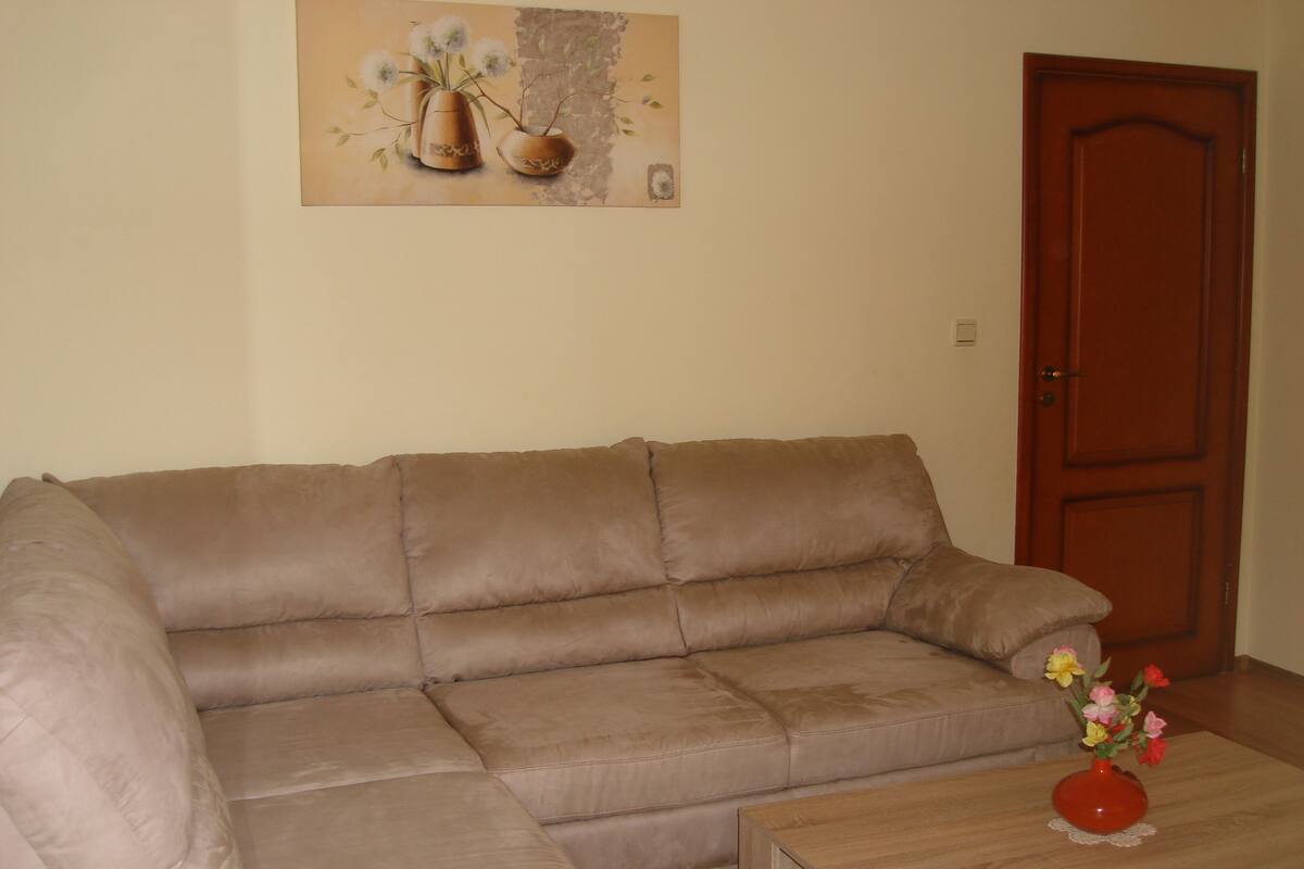 A living room with a beige sectional sofa, a wooden coffee table with a red vase, and a door in the background.