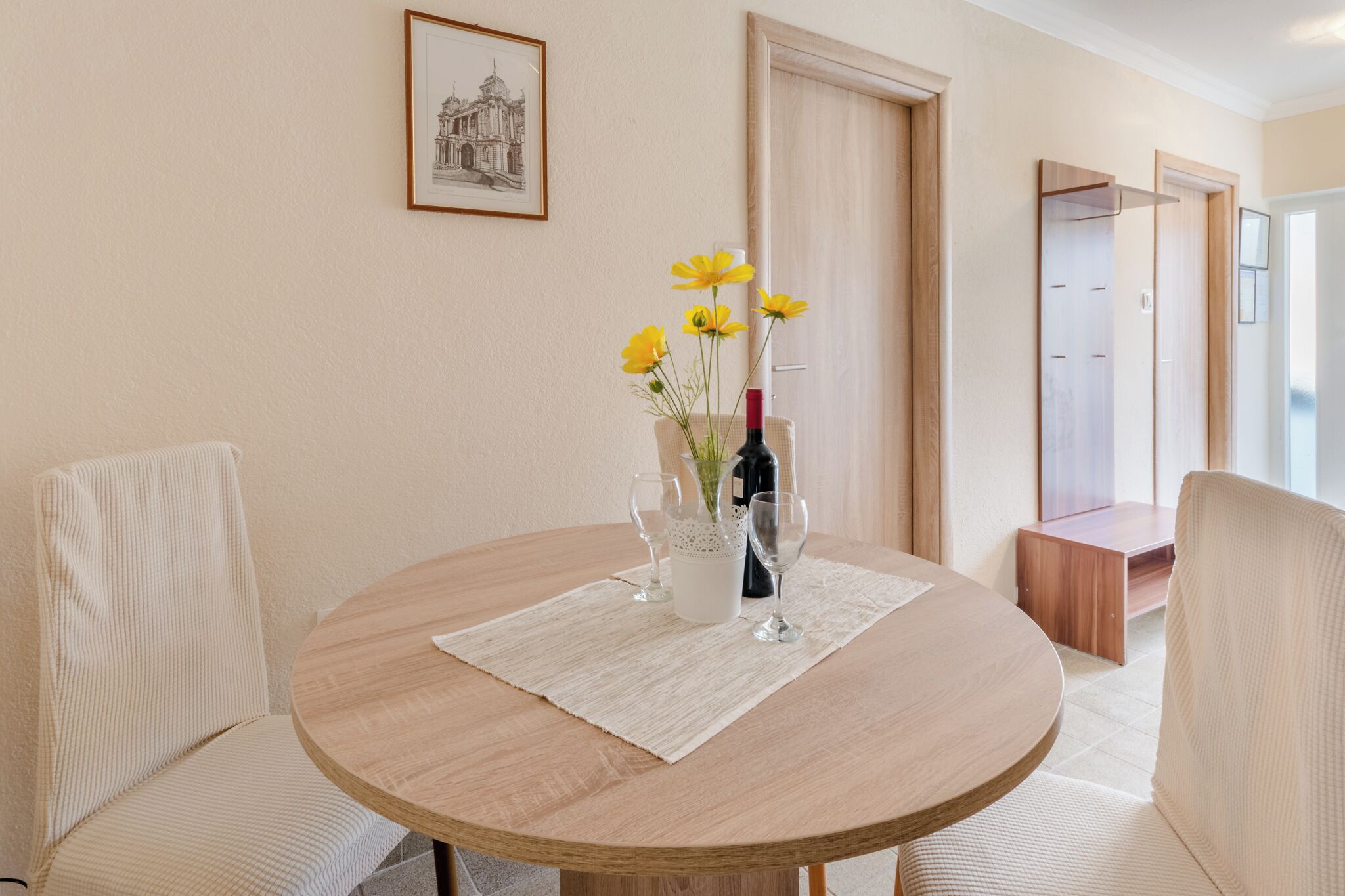A round table with a vase of yellow flowers, two wine glasses, and a bottle of wine. There are two upholstered chairs around the table. A framed picture hangs on the wall above the table.
