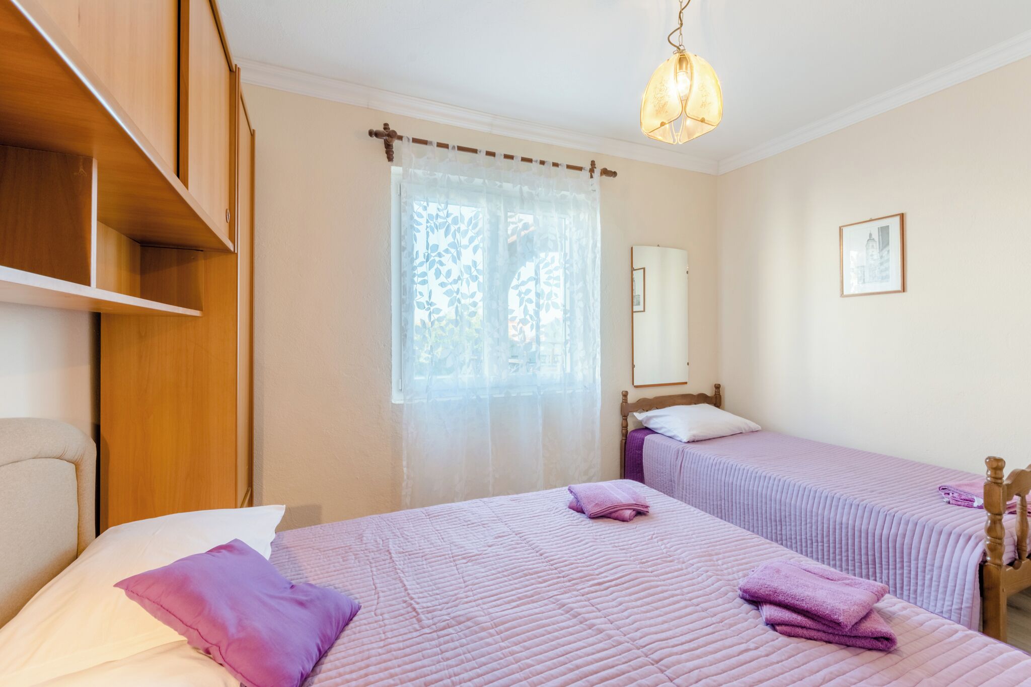 A bedroom with two beds, wooden headboards, a large window with sheer curtains, and a hanging light fixture.