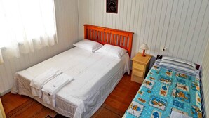 Ein Einzelbett mit weißer Bettwäsche, ein Holz-Kopfbrett, ein Nachttisch mit Lampe und ein Fenster mit durchsichtigen Vorhängen.