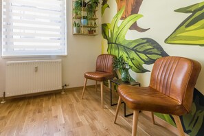 A room with two leather chairs, a radiator, a window with blinds, and a wall with a large plant mural. - Urban Jungle Designer Apartment (Mannheim)