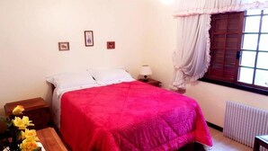 A bedroom with a bed, a nightstand, a lamp, a window with curtains, and a radiator. - Apartamento da Borges (Gramado)