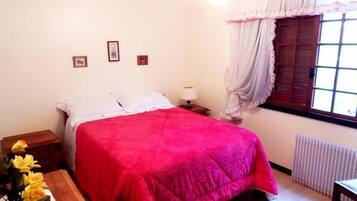 A bedroom with a bed, a nightstand, a lamp, a window with curtains, and a radiator.