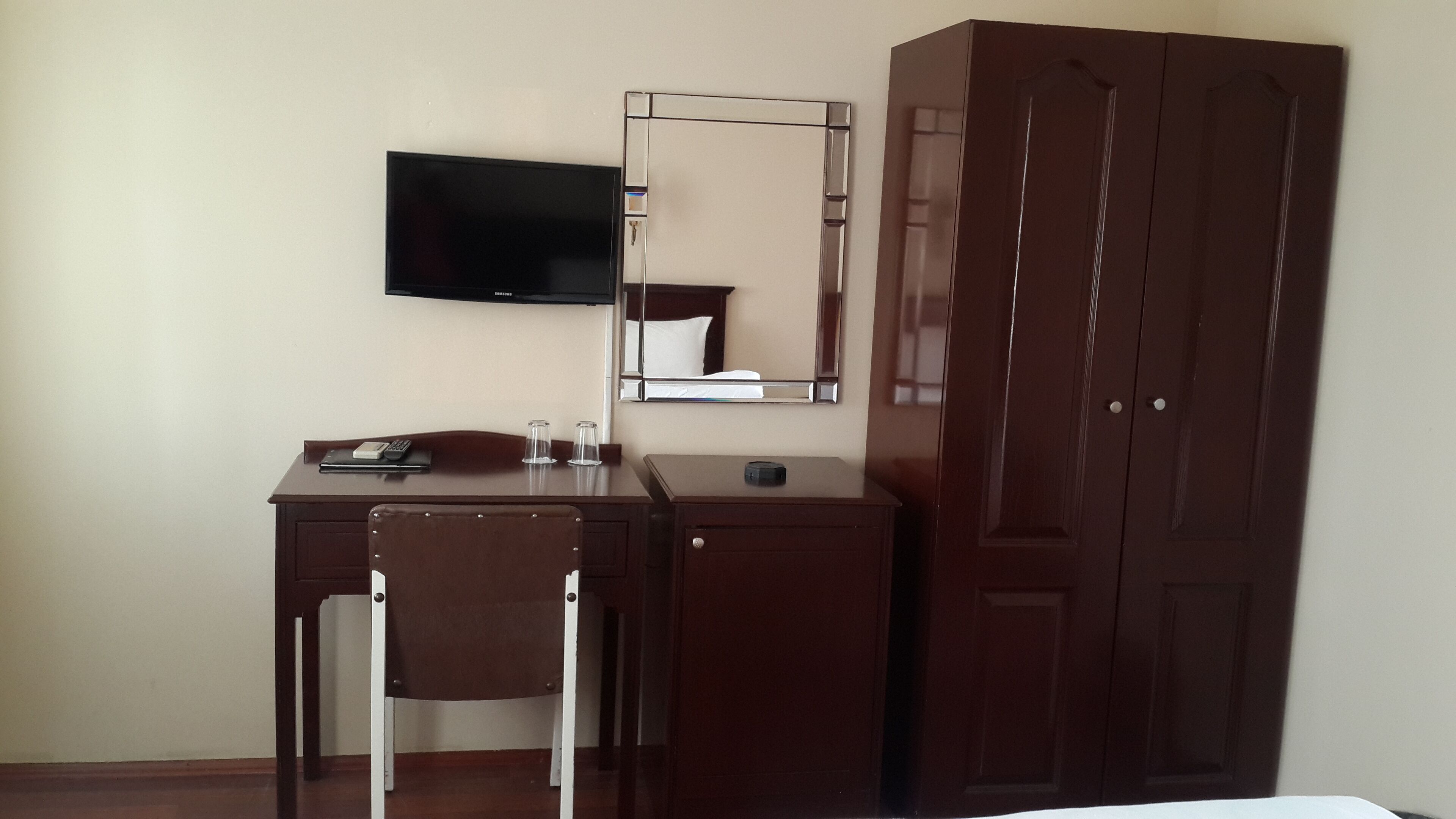 A hotel room with a wooden desk, a chair, a flat-screen TV mounted on the wall, a mirror, and a wardrobe.