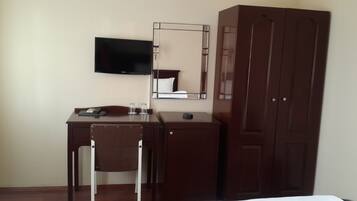 A hotel room with a wooden desk, a chair, a flat-screen TV mounted on the wall, a mirror, and a wardrobe.