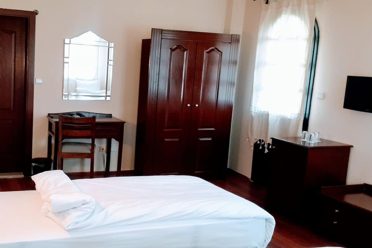 Quarto de hotel com cama, escrivaninha, cadeira, guarda-roupa, televisão e janela com cortinas.