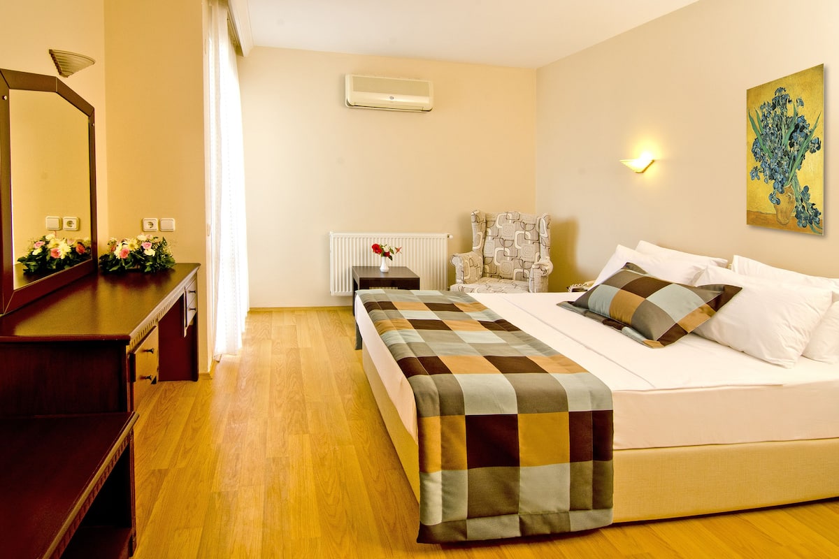 Habitación de hotel con piso de madera, cama con colcha de cuadros, mesita de noche con un jarrón de flores, una silla, aire acondicionado instalado en la pared y un cuadro en la pared.