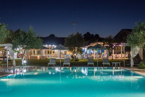 Piscina con sillas de descanso y una zona cubierta, rodeada de árboles y edificios con iluminación exterior.