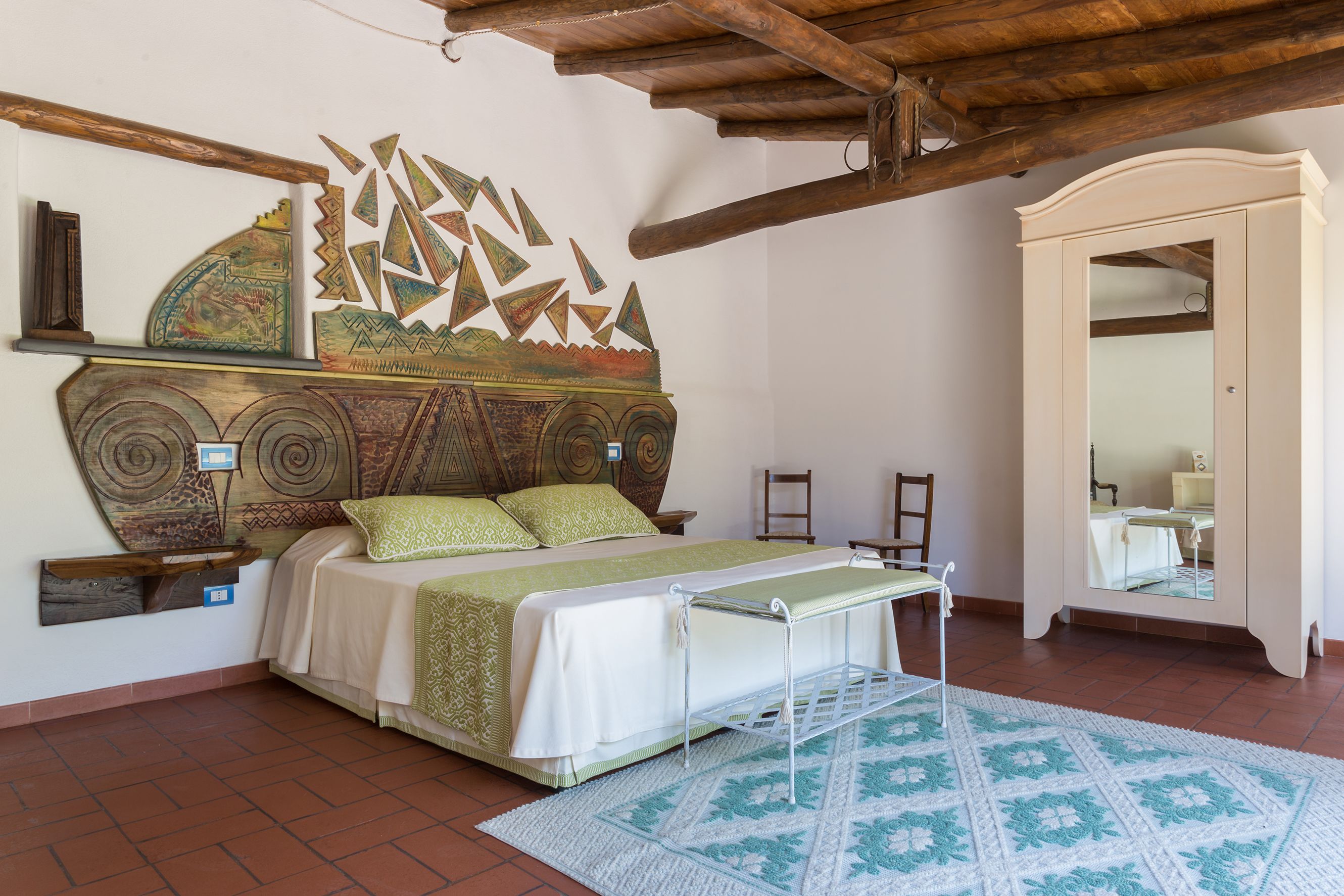 Un dormitorio con cama, cabecera decorativa, techo de madera y suelo de baldosas.