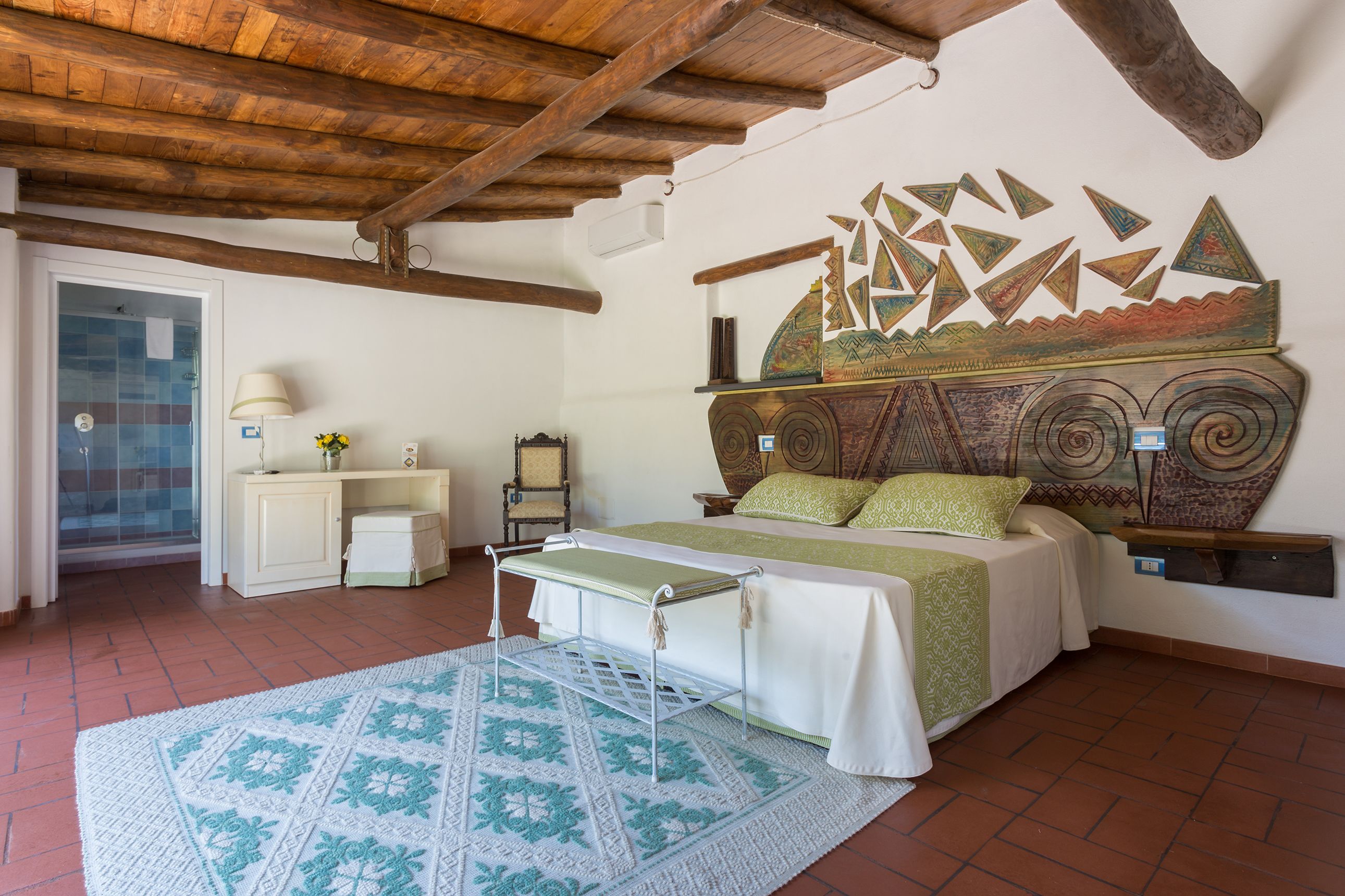 A bedroom with a wooden ceiling, a bed with green bedding, a patterned rug, and a wall with a decorative sailboat painting.
