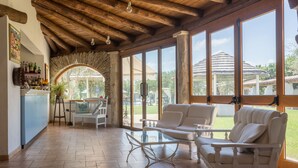 A spacious living area with a wooden ceiling, large windows, a white sofa, a glass coffee table, and a bar area with bottles and a plant. - Da Renzo (Siamaggiore)