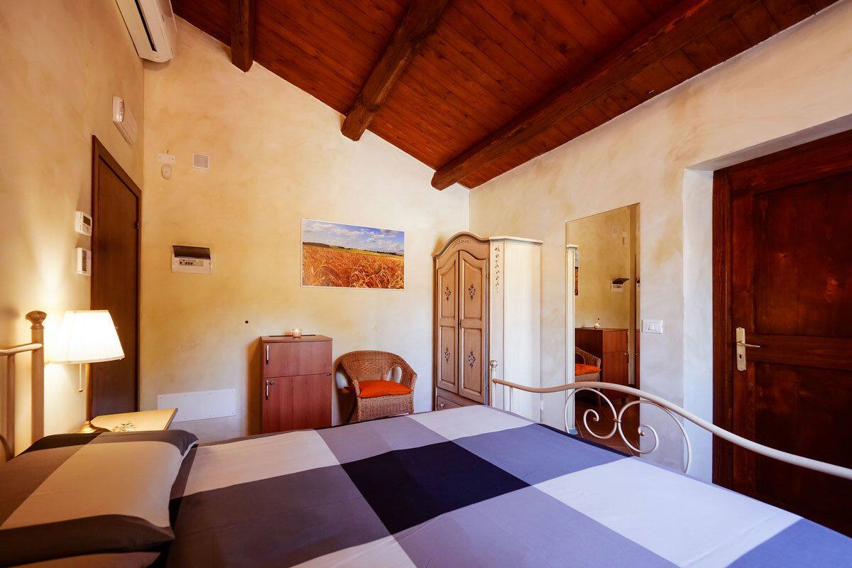 Une chambre avec une couvre-lit à carreaux, un plafond en bois, une chaise en osier, une armoire et une porte.