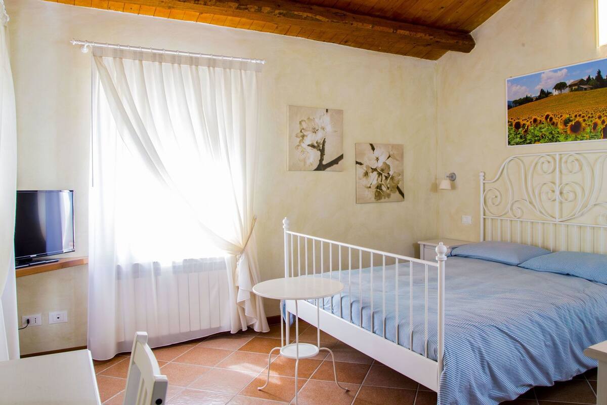 Une chambre à coucher avec un lit, une télévision, une petite table et une fenêtre avec des rideaux.